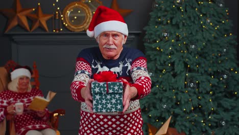 Mature-grandfather-in-New-Year-sweater-and-hat-presenting-Christmas-gift-box,-holidays-at-home