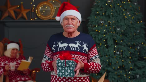 Elderly-grandfather-in-festive-sweater-presenting-Christmas-gift-box,-smiling,-looking-at-camera