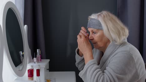 Old-senior-elderly-woman-grandmother-applying-anti-wrinkle-eye-patch,-putting-makeup-on-at-home