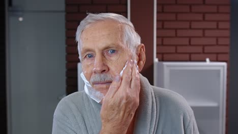 Senior-man-grandfather-in-bathrobe-shaving-with-manual-shaving-blade,-looking-into-mirror.-POV-shot