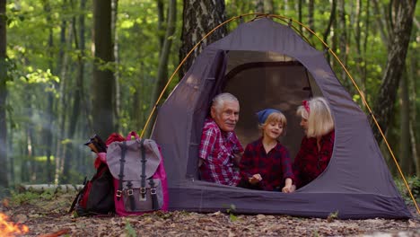 Senior-grandmother-grandfather-with-granddaughter-sitting-in-tourist-tent-over-campfire-in-wood