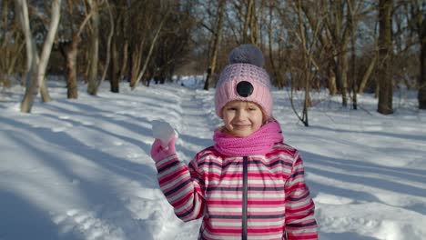 Child-girl-throwing-snowball-towards-camera,-smiling-kid-walking,-playing-with-snow-in-winter-park