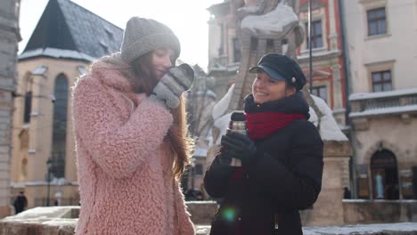 Two-smiling-women-tourists-traveling-together,-drinking-hot-tea,-coffee-from-thermos-on-city-street