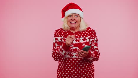Großmutter-Im-Weihnachtspullover-Schaut-Auf-Smartphone-Display-Und-Freut-Sich-Aufrichtig-über-Erfolg-Und-Glück