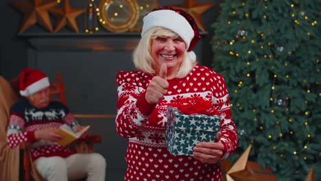 Positive-senior-grandmother-in-Christmas-sweater-smiling-friendly-at-camera-and-raises-thumbs-up