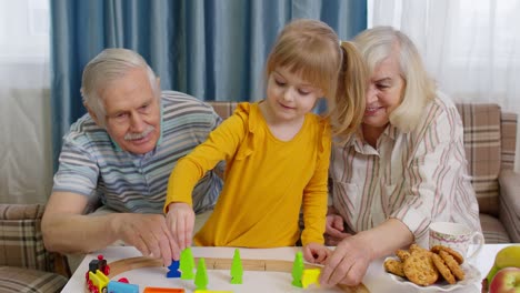 Mature-couple-grandmother,-grandfather-with-child-girl-grandkid-riding-toy-train-on-railroad-at-home