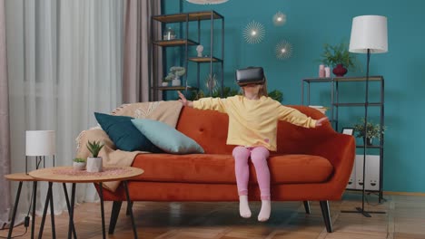 Toddler-girl-sitting-on-home-sofa-using-virtual-reality-headset-helmet-app-to-play-simulation-game