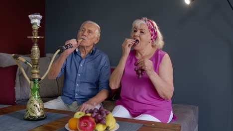 Stylish-senior-couple-enjoy-smoking-hookah,-eating-fruits-at-home,-celebrating,-having-fun-together