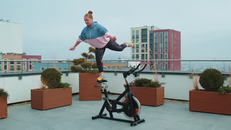 Athletic-girl-performing-swallow-acrobatic-trick-exercises-on-cycling-stationary-bike-on-house-roof