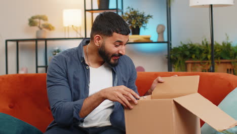 Indian-man-unpacking-delivery-parcel-receiving-purchase-by-postal-shipping-courier-sitting-on-sofa