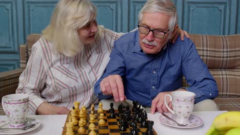 Smiling-senior-couple-grandfather-grandmother-resting-on-sofa-drinking-coffee,-playing-chess-at-home