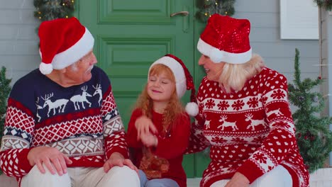 Senior-grandparents-with-granddaughter-in-Santa-Claus-hat-celebrating-Christmas-near-decorated-house
