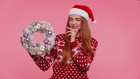 Adult-smiling-girl-in-red-Christmas-holiday-sweater-hold-raise-point-finger-on-festive-wreath-toy