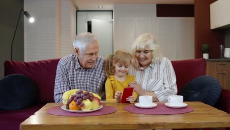 Senior-Caucasian-couple-with-cute-child-girl-granddaughter-using-mobile-phone,-watching-funny-videos