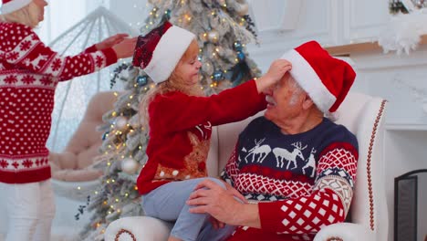 Granddaughter-child-fixes-Christmas-Santa-Claus-hat-on-happy-senior-old-smiling-grandfather-at-home