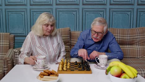 Smiling-senior-couple-grandfather-grandmother-resting-on-sofa-drinking-coffee,-playing-chess-at-home