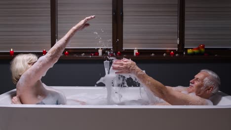 Senior-couple-grandfather-and-grandmother-is-taking-bath,-splashing-water-in-bathroom-with-candles