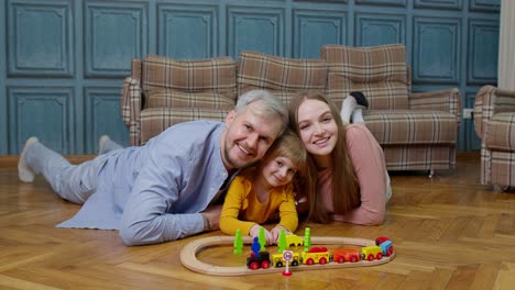 Young-mother-and-father-playing-with-child-daughter-riding-toy-train-on-wooden-railroad-game-at-home