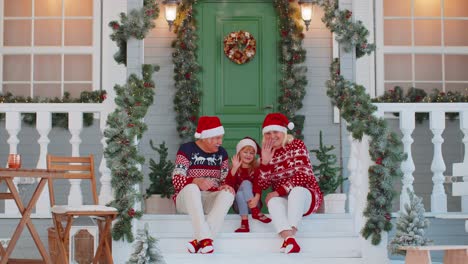 Senior-grandmother-grandfather-with-granddaughter-sitting-at-Christmas-house-porch-waving-hello-hi