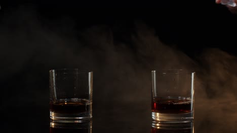 Barmans-dropping-ice-cubes-into-drinking-glasses-with-whiskey,-cognac,-brandy-on-black-background