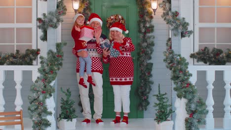Senior-grandmother-grandfather-with-granddaughter-standing-at-Christmas-house-porch-waving-hello-hi