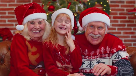 Senior-grandparents-with-granddaughter-in-Santa-hats-laughing-out-loud-at-home-near-Christmas-tree