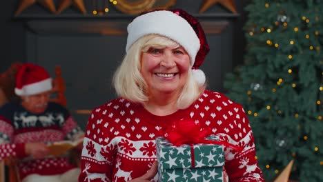 Ältere-Großmutter-Im-Festlichen-Pullover-Präsentiert-Weihnachtsgeschenkbox,-Lächelt,-Blickt-In-Die-Kamera
