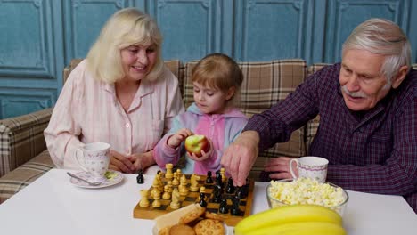 Mature-grandmother-grandfather-with-child-girl-grandchild-playing-chess-game-with-on-table-in-room