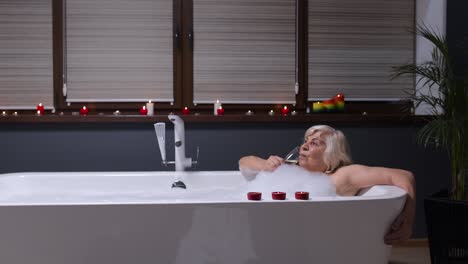 Active-senior-woman-lying-in-warm-bath-with-bubbles,-drinking-champagne-after-hard-working-day