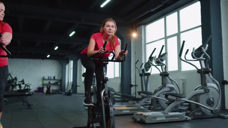 Group-class-of-two-friends-women-exercising,-training,-spinning-on-stationary-bike-at-modern-gym