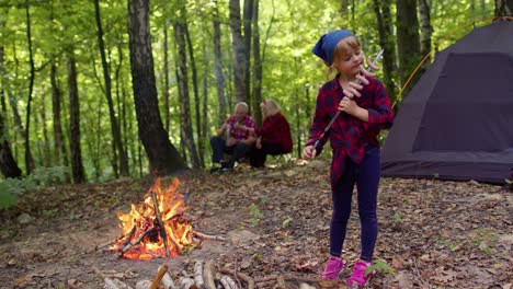 Child-girl-eating-cooked-fried-sausages-near-campfire,-senior-grandparents-on-background-in-wood