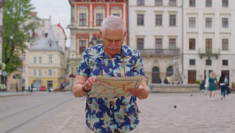 Elderly-stylish-tourist-grandfather-man-walking-along-street-looking-for-way-using-paper-map-in-city
