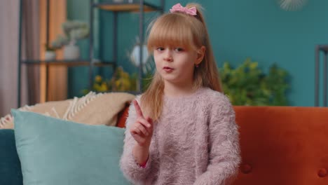 Child-girl-kid-sitting-on-sofa-at-home-alone-say-no-hold-palm-folded-crossed-hands-in-stop-gesture