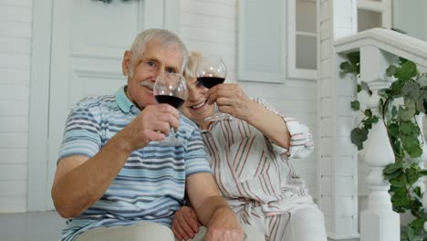 Attractive-senior-elderly-Caucasian-couple-sitting-and-drinking-wine-in-porch-at-home,-making-a-kiss