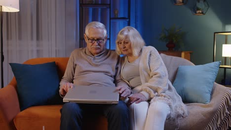 Feliz-Pareja-De-Ancianos-Usando-Una-Computadora-Portátil-Hablando-Juntos-Haciendo-Compras-En-Línea-En-El-Sofá-De-Casa