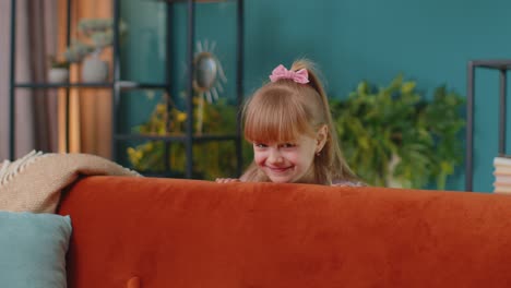 Child-girl-playing-hide-and-seek-peekaboo-game-near-sofa-at-home-alone-looking-at-camera-and-smiling
