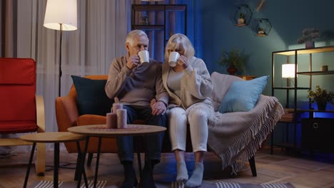Carefree-mature-family-couple-relaxing-on-sofa-at-home,-enjoying-conversation-with-cups-of-hot-tea