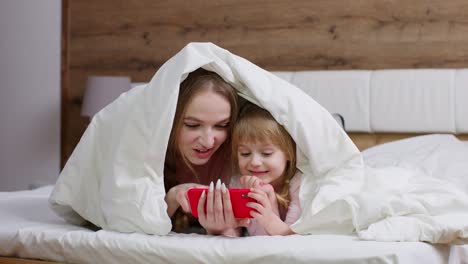 Mother-lying-on-bed-under-duvet-blanket-with-daughter-kid-girl-holding-mobile-phone-watching-cartoon