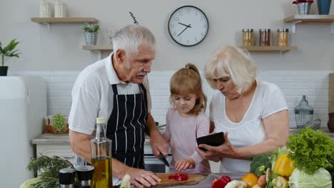 Senior-grandparents-couple-with-digital-tablet-and-granddaughter-cutting-vegetables-in-kitchen