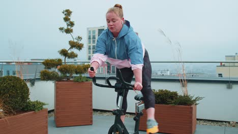 Healthy-Caucasian-woman-exercising-workout-on-stationary-cycling-machine-bike-on-house-rooftop-gym