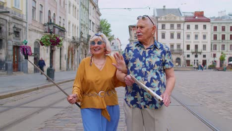 Senior-old-tourists-man-with-woman-walking-in-city-with-smartphone-on-selfie-stick-and-taking-photos