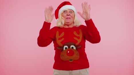 Grandmother-woman-in-Christmas-sweater-with-deers-waves-hand-palm-in-hello-gesture-welcomes-someone
