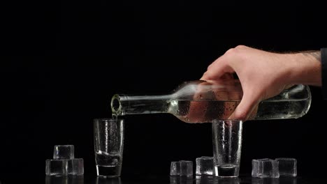 Bartender-pouring-up-vodka-from-bottle-into-two-shots-glasses-with-ice-cubes-on-black-background