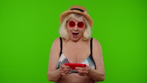 Senior-old-exited-woman-tourist-in-swimsuit-playing-video-game-on-mobile-cell-phone-on-chroma-key