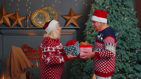 Romantic-senior-couple-presenting-gift-boxes-to-each-other-at-home-near-decorating-Christmas-tree