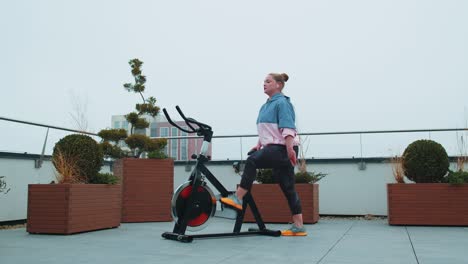 Healthy-caucasian-woman-exercising-workout-on-stationary-cycling-machine-bike-on-house-rooftop-gym