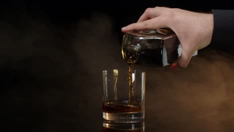 Barman-pour-whiskey-cognac-brandy-from-bottle-into-drinking-glass-with-ice-cubes-on-dark-background