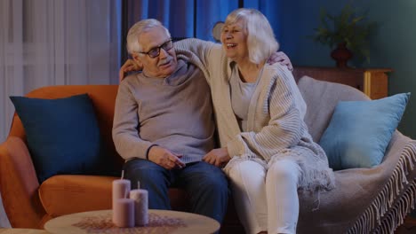 Happy-old-senior-elderly-family-couple-hugging,-laughing,-smiling-talking-at-modern-home-sofa