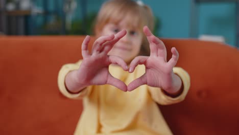Smiling-child-girl-kid-on-home-sofa-looking-at-camera-makes-heart-gesture-demonstrates-love-sign
