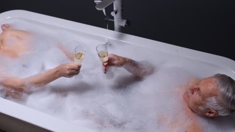 Senior-couple-man-and-woman-lying-in-warm-bath-with-bubbles,-enjoying-relaxation,-drinking-champagne